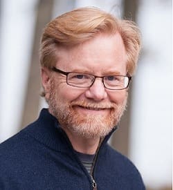 A person with glasses, a beard, and blonde hair, wearing a dark sweater, smiles at the camera.