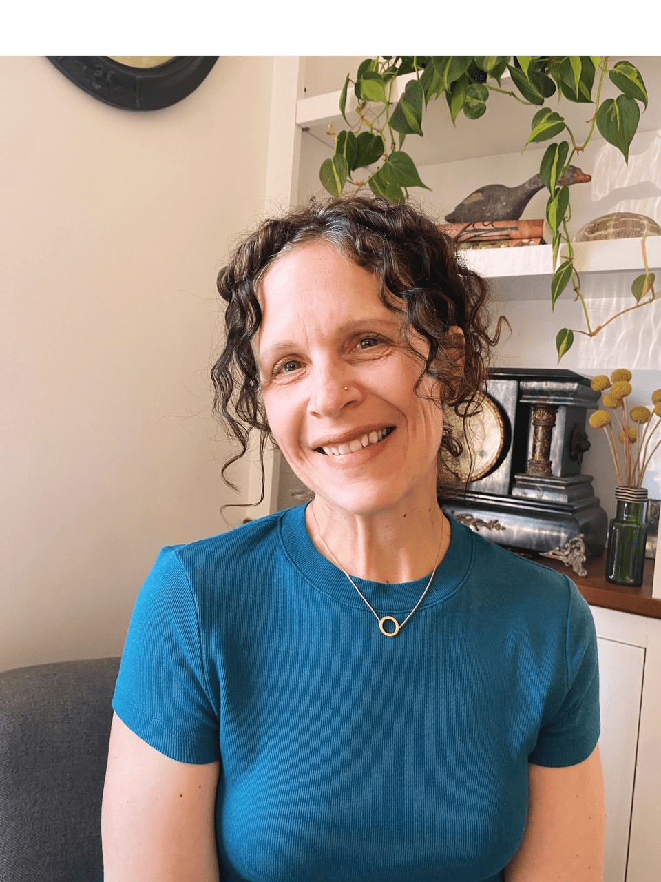 A person with curly hair smiles while seated indoors, wearing a teal shirt and necklace. The background features shelves with decor and a plant.