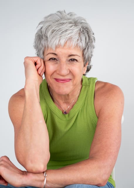 Person with short gray hair wearing a green sleeveless top, sitting with one arm resting on a knee, smiling at the camera against a neutral background.