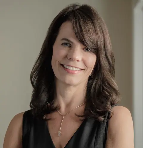 Kiera Preston A woman with shoulder-length brown hair wearing a black sleeveless top and a pendant necklace smiles at the camera in a neutral indoor setting.