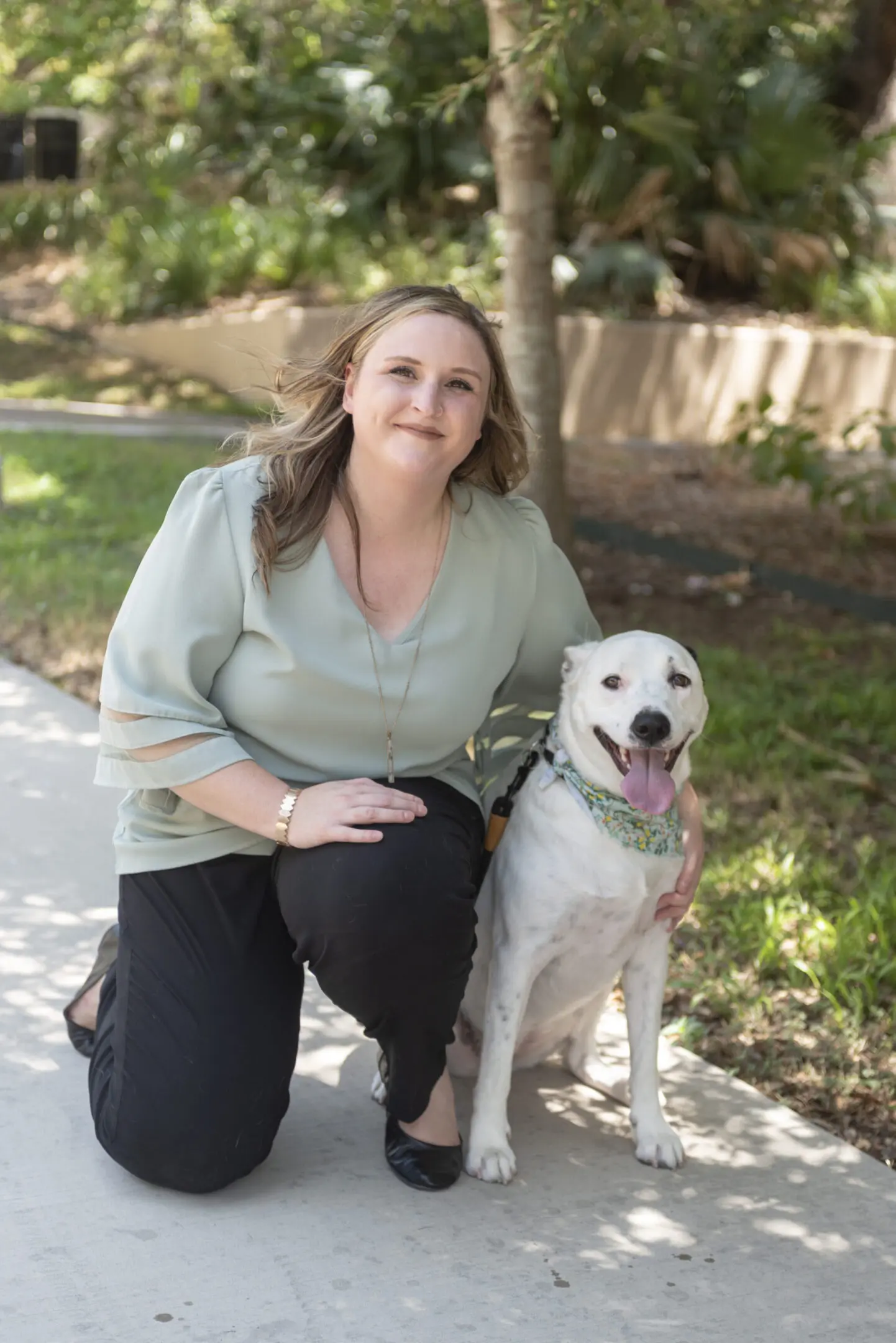 tiana Woman kneeling on a sidewalk next to a panting white dog wearing a bandana, with trees and grass in the background.
