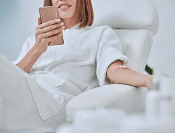 A woman in a white robe sits in a chair, looking at her smartphone while receiving an intravenous (IV) drip in her arm.