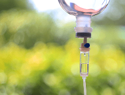 Close-up of an intravenous (IV) drip with fluid inside, hanging outdoors with a blurred green background.