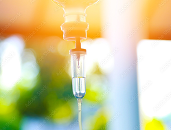 Close-up of an IV drip chamber with clear fluid inside, set against a blurred background with sunlight.