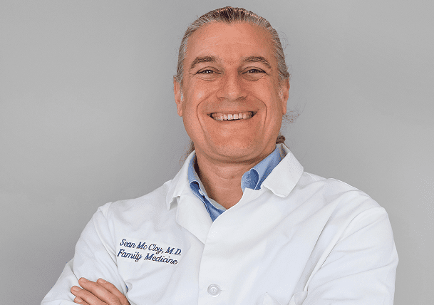 A smiling person with gray hair, wearing a white lab coat embroidered with “Sean McClay, M.D. Family Medicine,” stands against a plain light gray background.