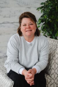 A woman with short brown hair sits on a patterned chair, wearing a light gray sweater and black pants, with her hands clasped and a slight smile on her face. A leafy plant is in the background.